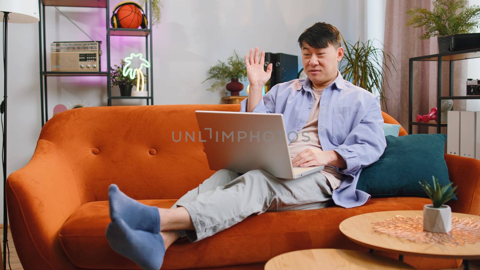 Asian man sitting on couch looking at camera, making video webcam conference call with friends or family, enjoying pleasant conversation. Chinese middle-aged guy laughing waving hello at home room