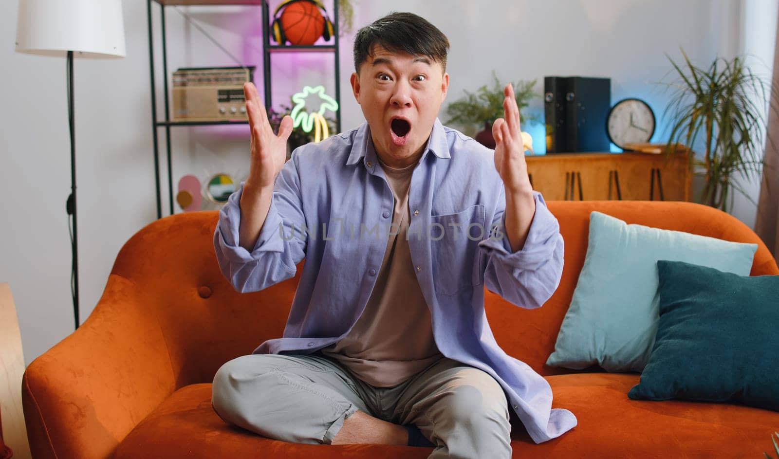 Happy asian man shouting wow, celebrating success, winning, goal achievement good news raising hands in gesture I did it, lottery luck at home apartment. Chinese guy in living room sitting on couch