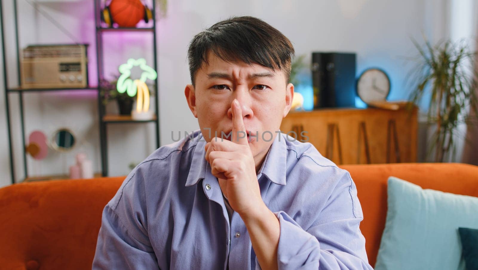 Shh be quiet, hush please. Asian man with presses index finger to lips makes silence hush gesture sign do not tells gossip secret. Chinese middle-aged guy at modern home living room sitting on couch