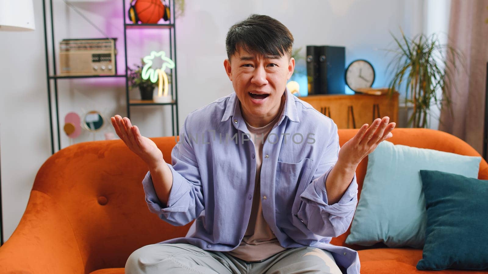 What. Why. Asian man raising hands in indignant expression asking reason of failure demonstrating disbelief irritation by troubles at modern home apartment. Chinese guy in living room sitting on sofa