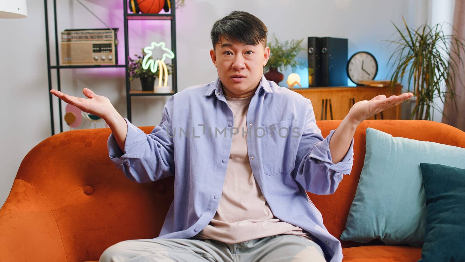 I dont know what to say. Confused asian handsome man feeling embarrassed about ambiguous question, having doubts, no idea, being clueless and uncertain at home. Chinese guy sits on sofa in living room
