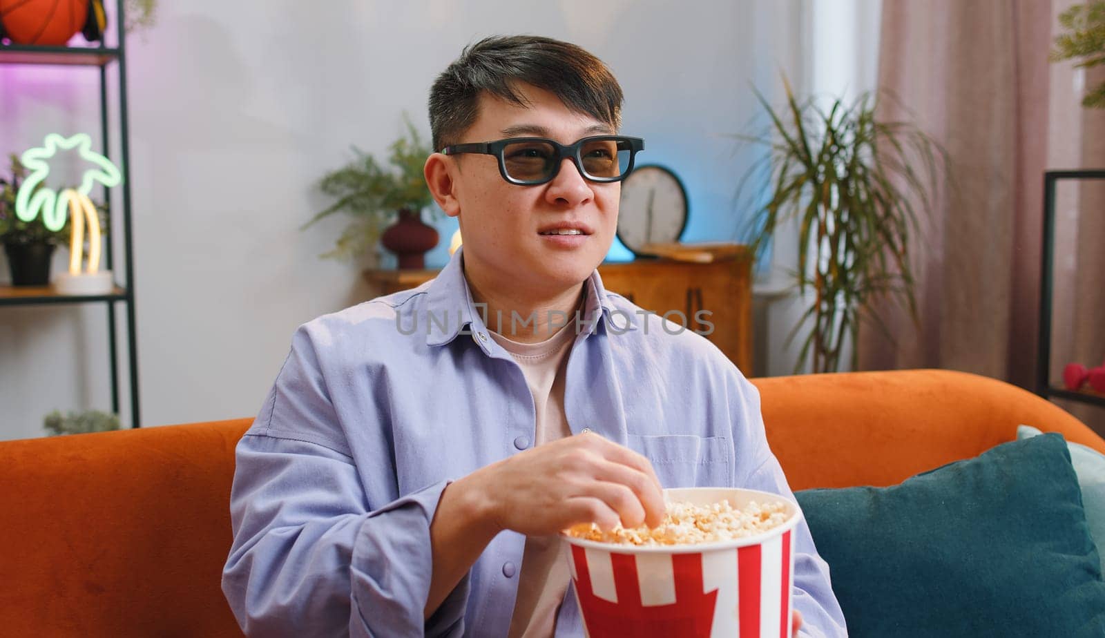 Excited asian man in 3D glasses sits on couch eating popcorn snacks and watching interesting TV serial sport game, film online social media movie content at home apartment. Guy enjoying entertainment
