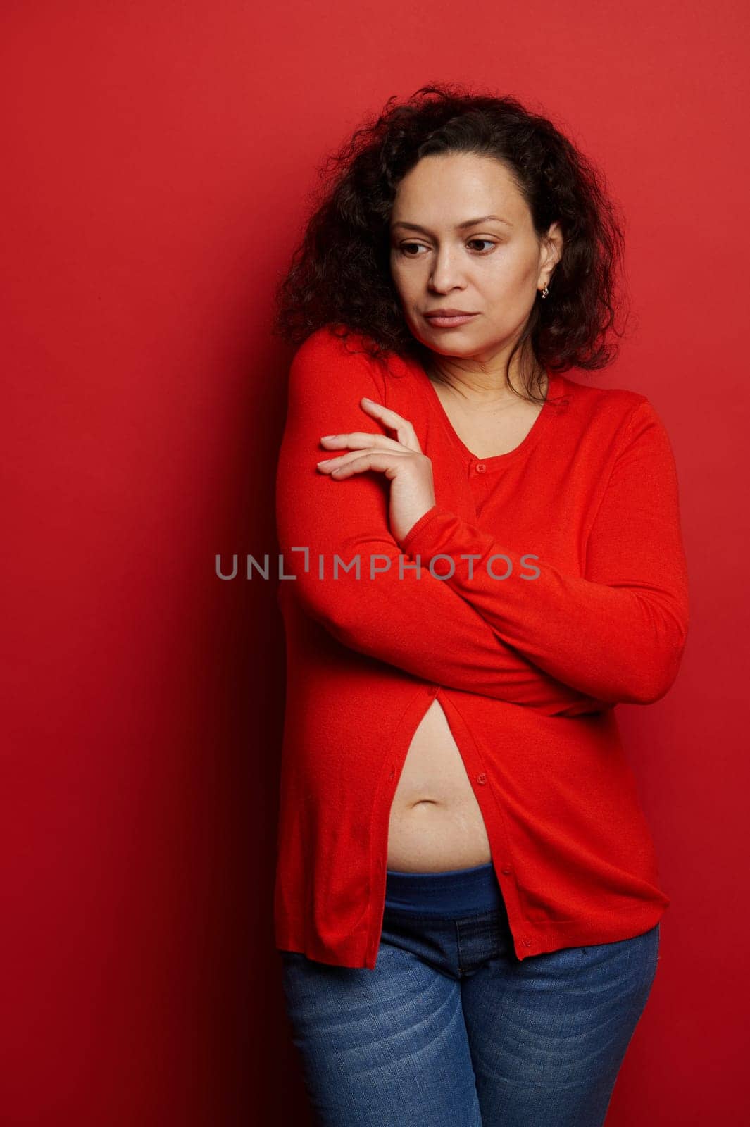 Pensive multi ethnic adult pregnant woman looking serious, posing with folded arms over red color background. Vertical studio portrait of a gravid female, expectant mother. Pregnancy 24 week.
