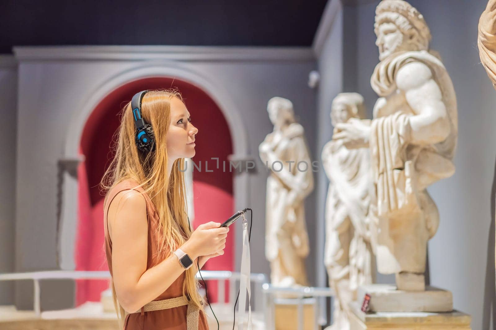 Portrait of contemporary young woman looking at sculptures and listening to audio guide at museum exhibition.