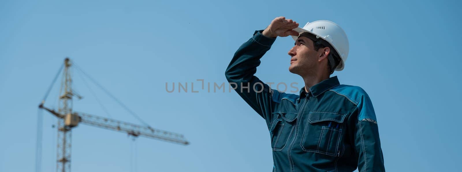 A builder in work clothes and a helmet stands on a construction site against the background of a construction crane