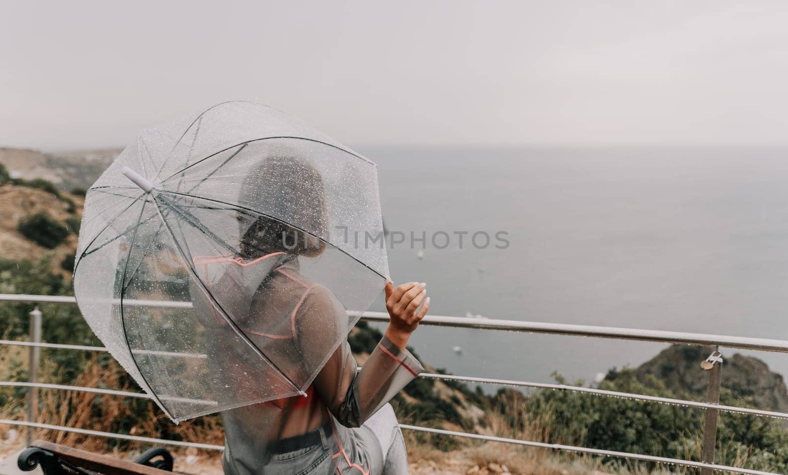 Woman rain umbrella. Happy woman portrait wearing a raincoat with transparent umbrella outdoors on rainy day in park near sea. Girl on the nature on rainy overcast day. Back view by panophotograph