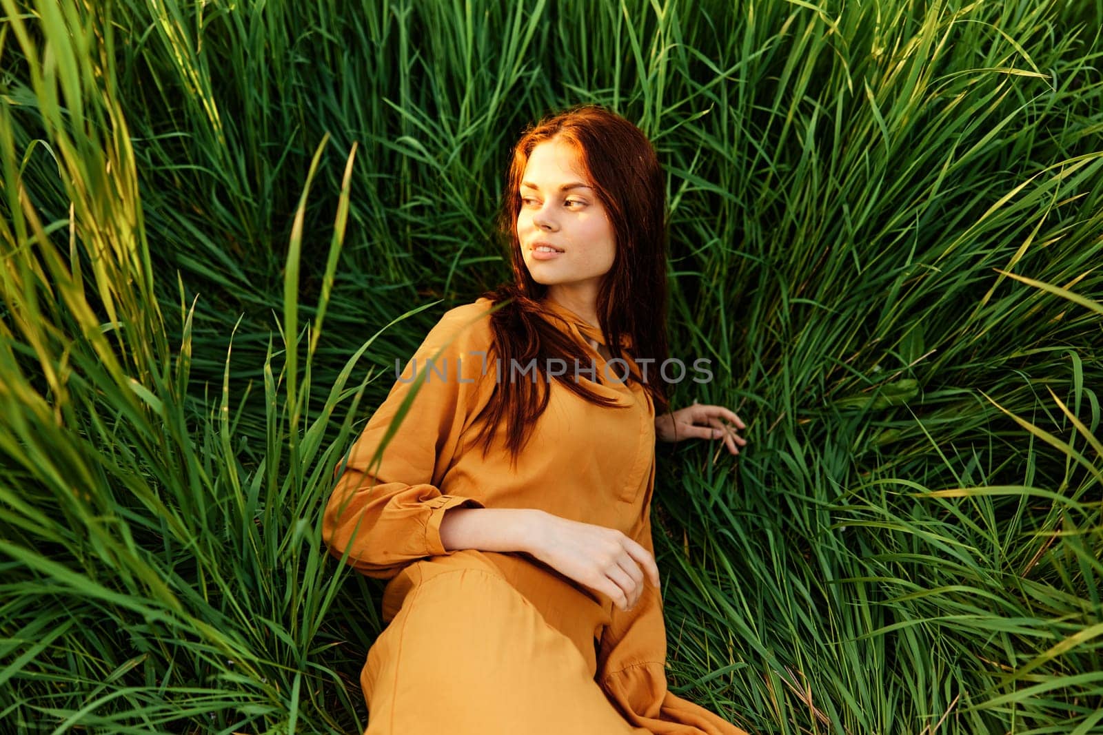 a happy woman lies in the tall green grass in a long orange dress and looks away smiling pleasantly. High quality photo