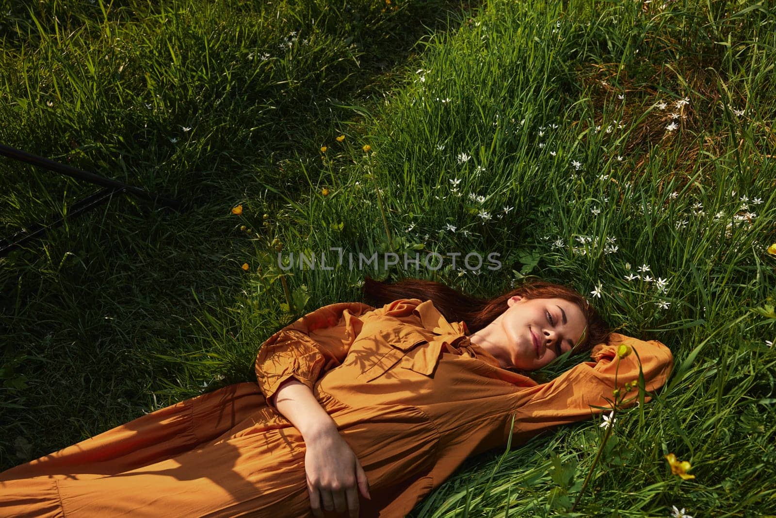 horizontal photo of a happy woman lying in the grass in a long orange dress, with her eyes closed and a pleasant smile on her face, with her hand behind her head. by Vichizh