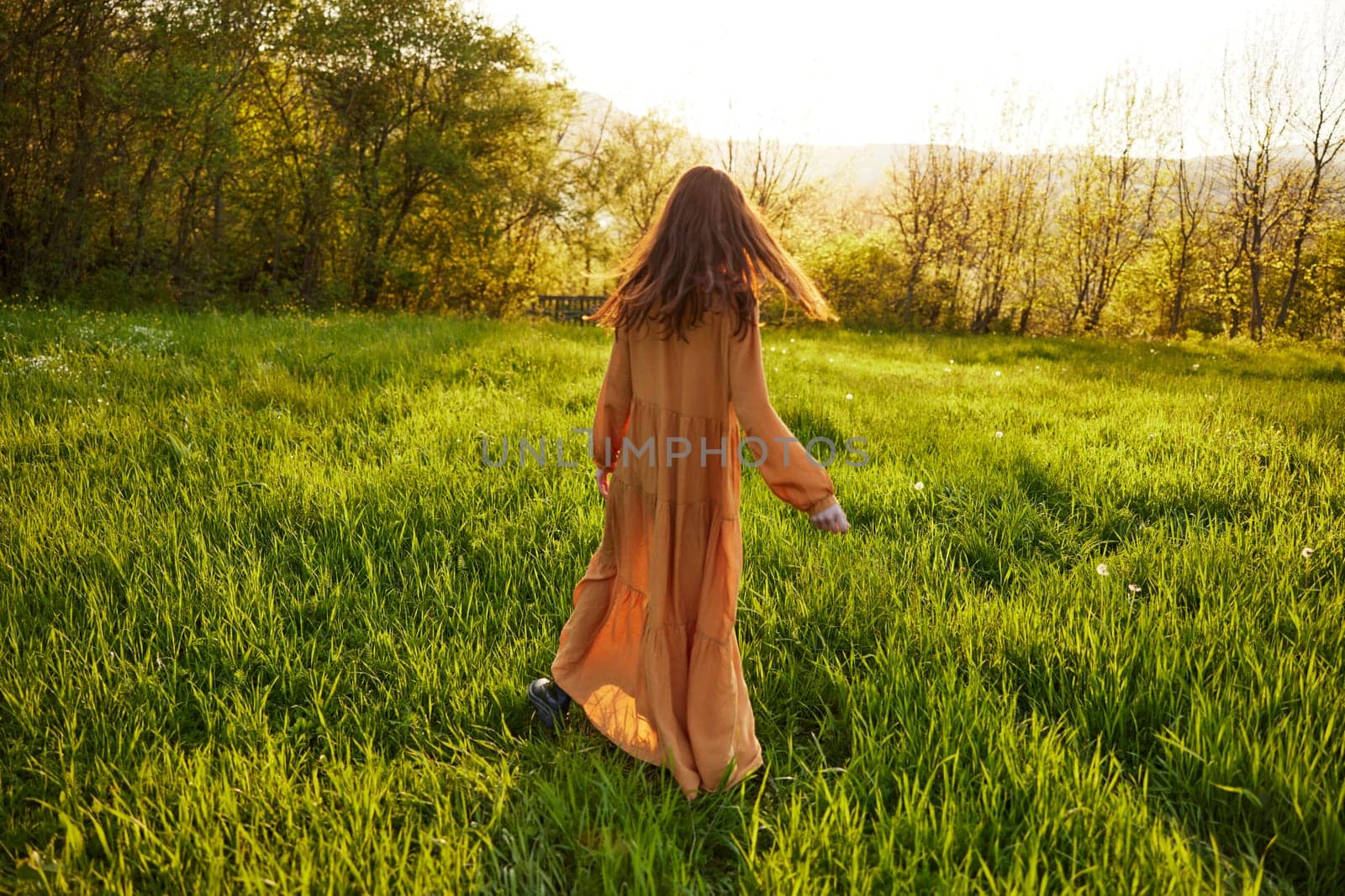 an attractive, slender, red-haired woman walks through a field during sunset, in a long orange dress enjoying unity with nature and relaxation holding the edge of her clothes. High quality photo