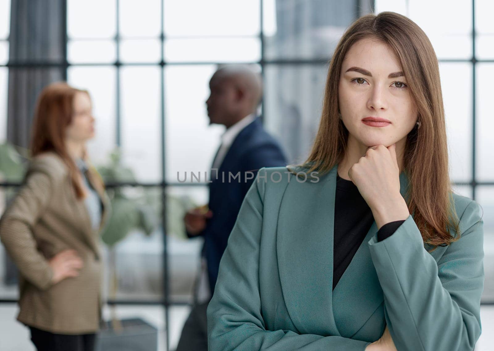 businesswoman standing with her arms crossed in an office while her colleagues have a meeting in the background.