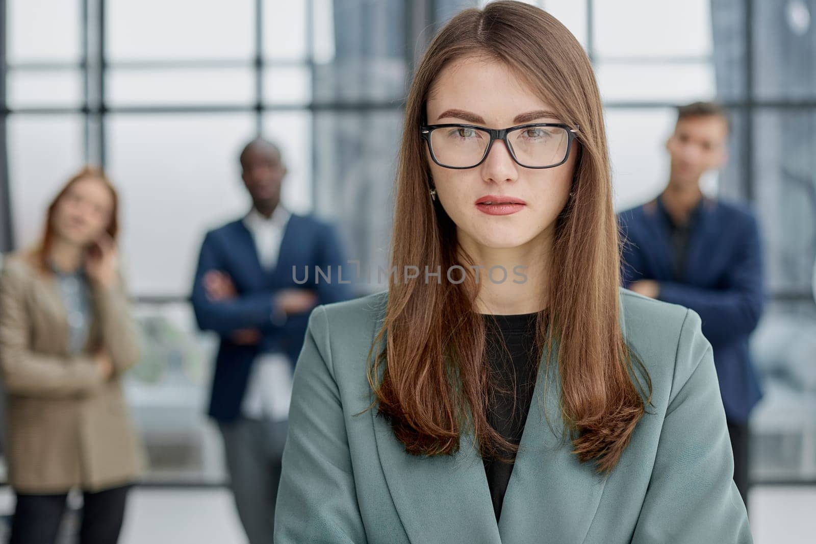 businesswoman standing with her arms crossed in an office while her colleagues have a meeting in the background.