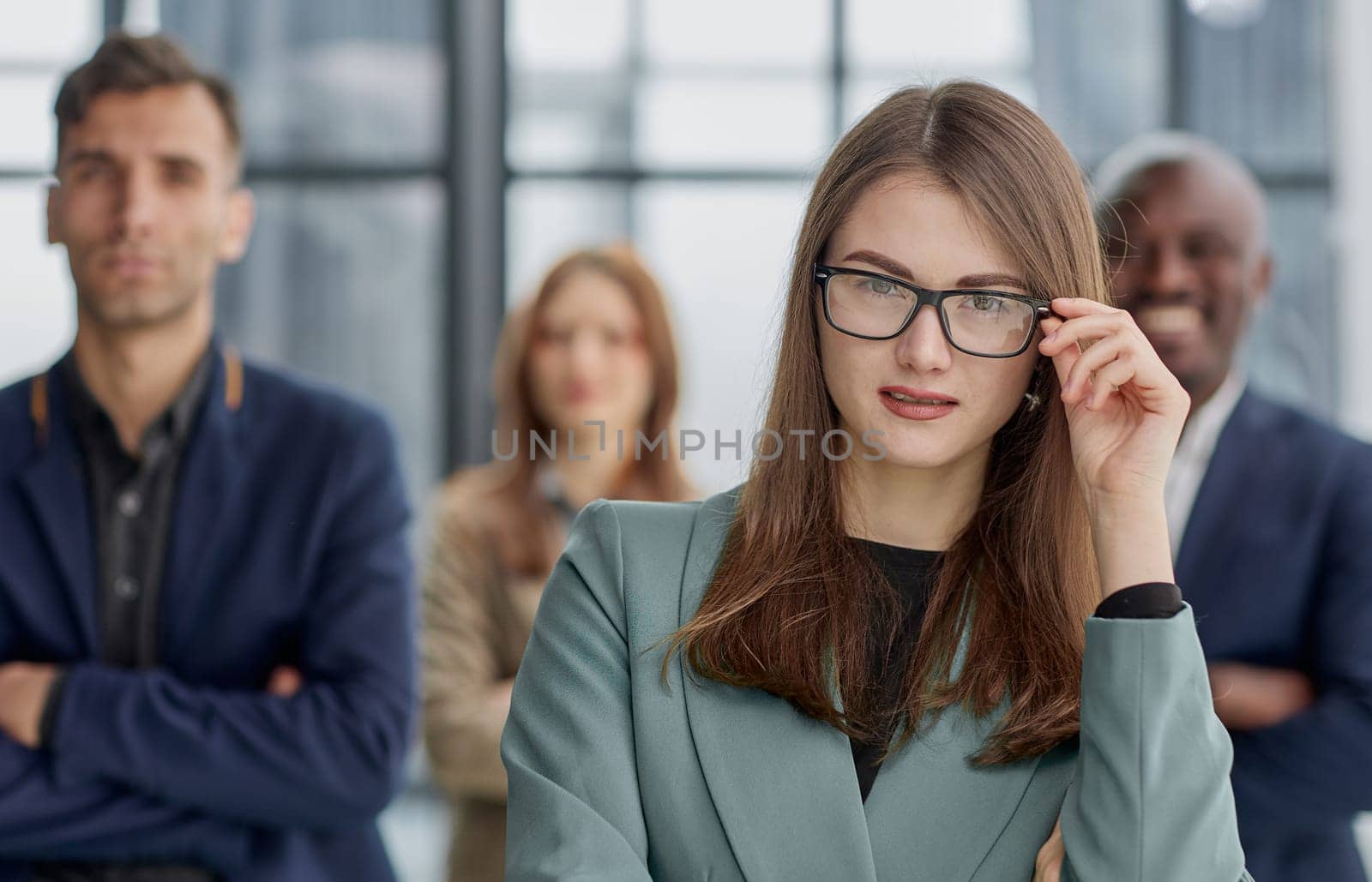 businesswoman standing with her arms crossed in an office while her colleagues have a meeting in the background.