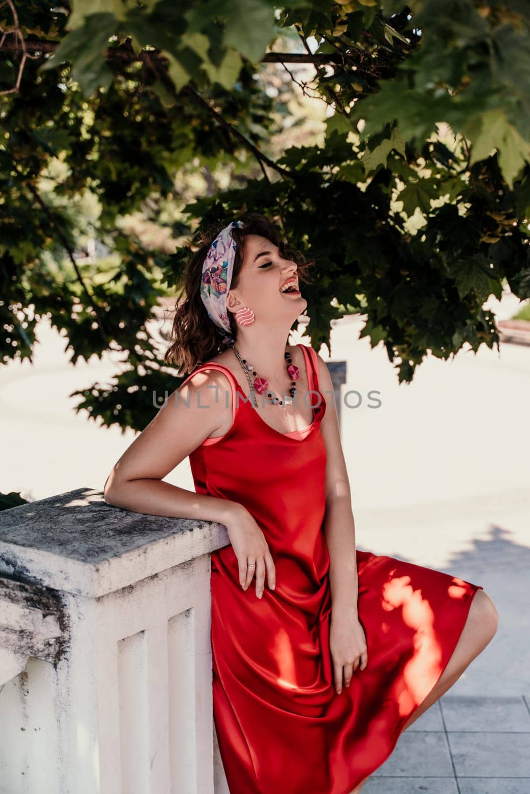 woman in a red silk dress and a bandage on her head smiles against the background of the leaves of a tree. She is leaning on the coop and looking into the camera. Vertical photo