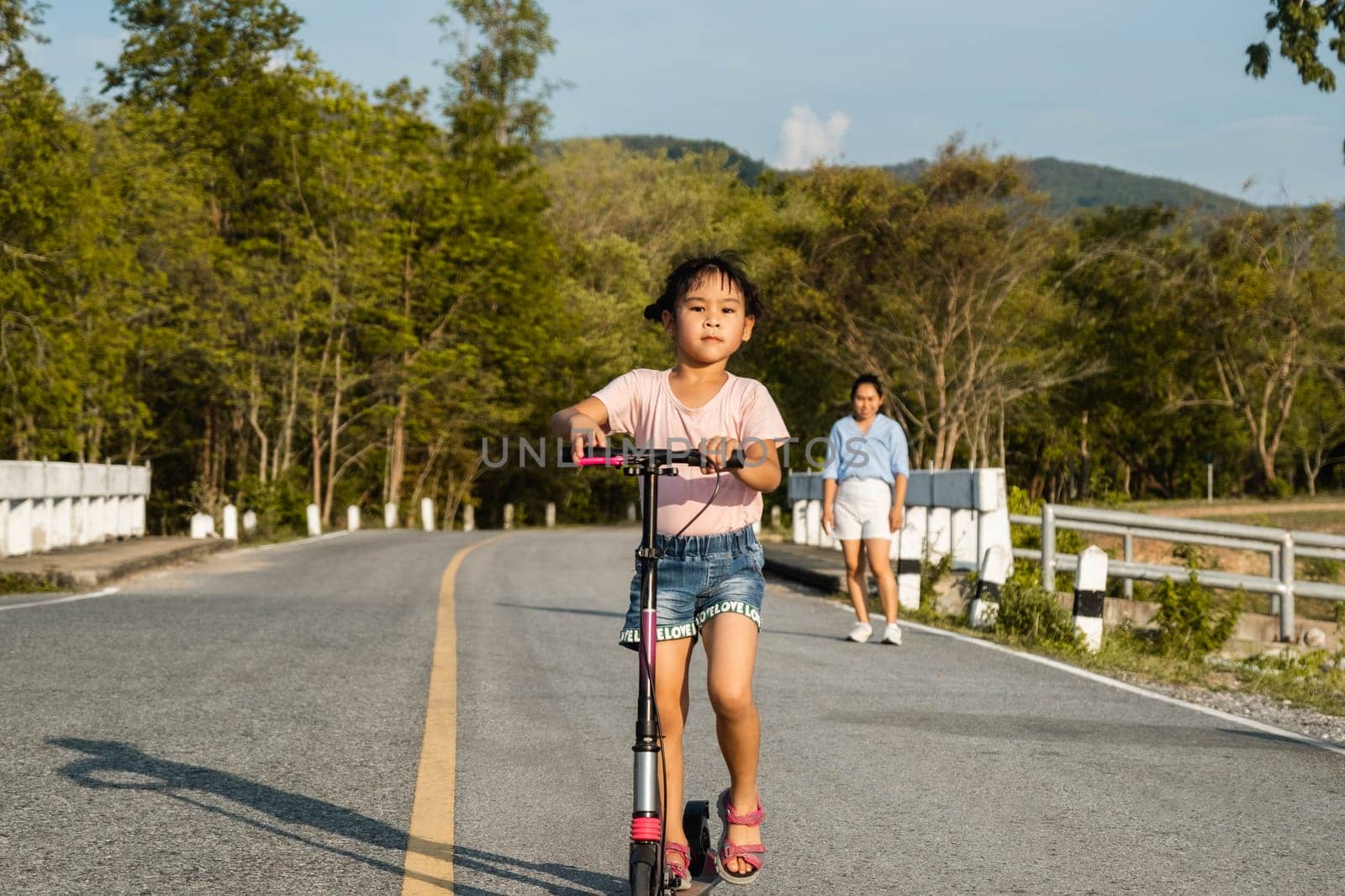 Cute little girl riding scooter on road in outdoor park. Healthy sports and outdoor activities for school children in the summer. by TEERASAK
