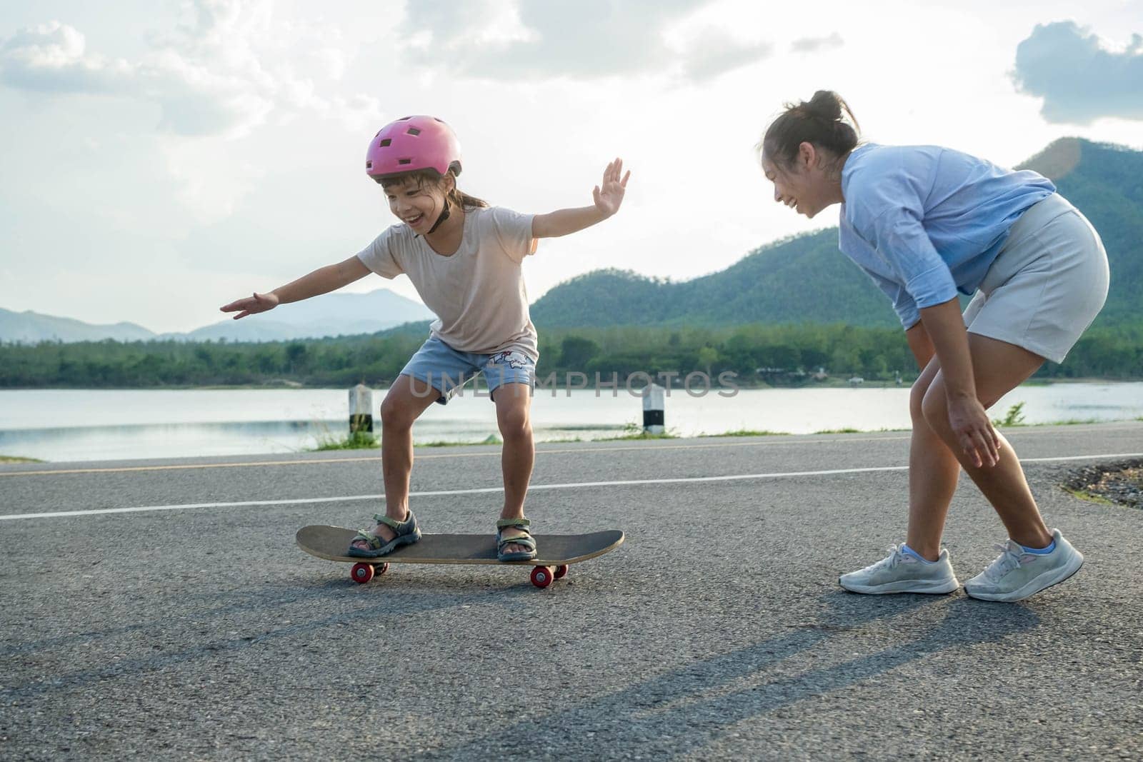 Mother teaching her daughter how to skateboard in the park. Child riding skate board. Healthy sports and outdoor activities for school children in the summer. by TEERASAK