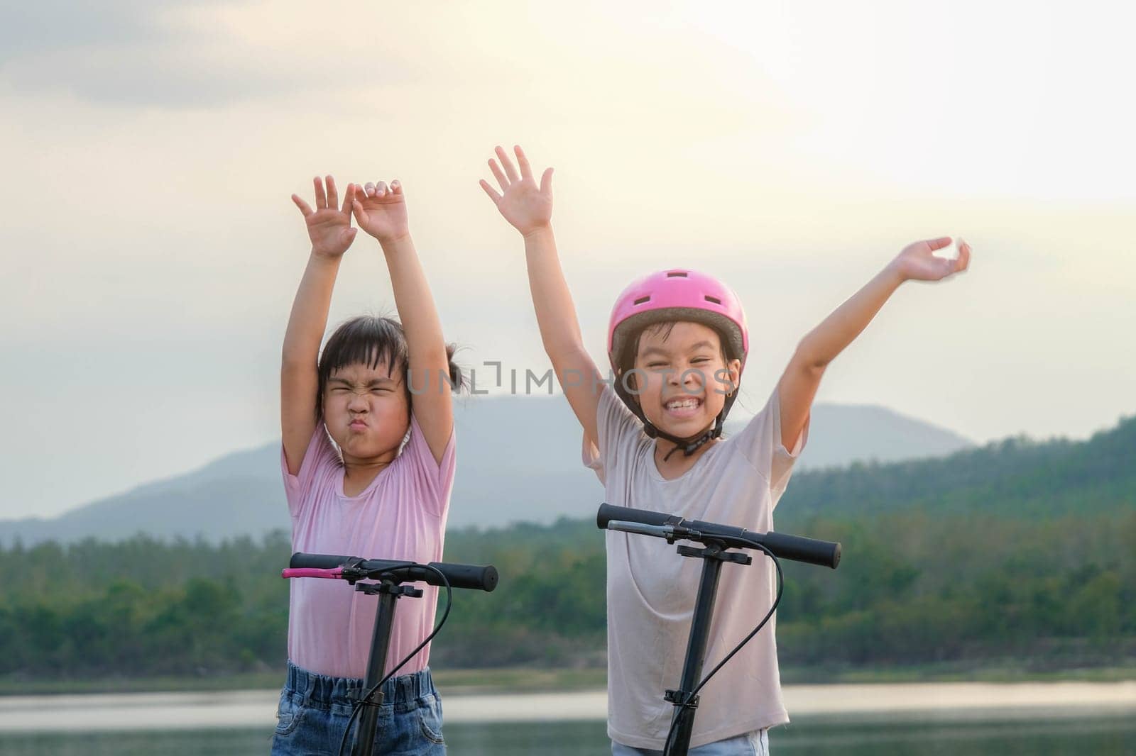 Two cute little girls smiling and posing together in summer garden. Happy kid riding kick scooter in the park. Healthy sports and outdoor activities for children. by TEERASAK