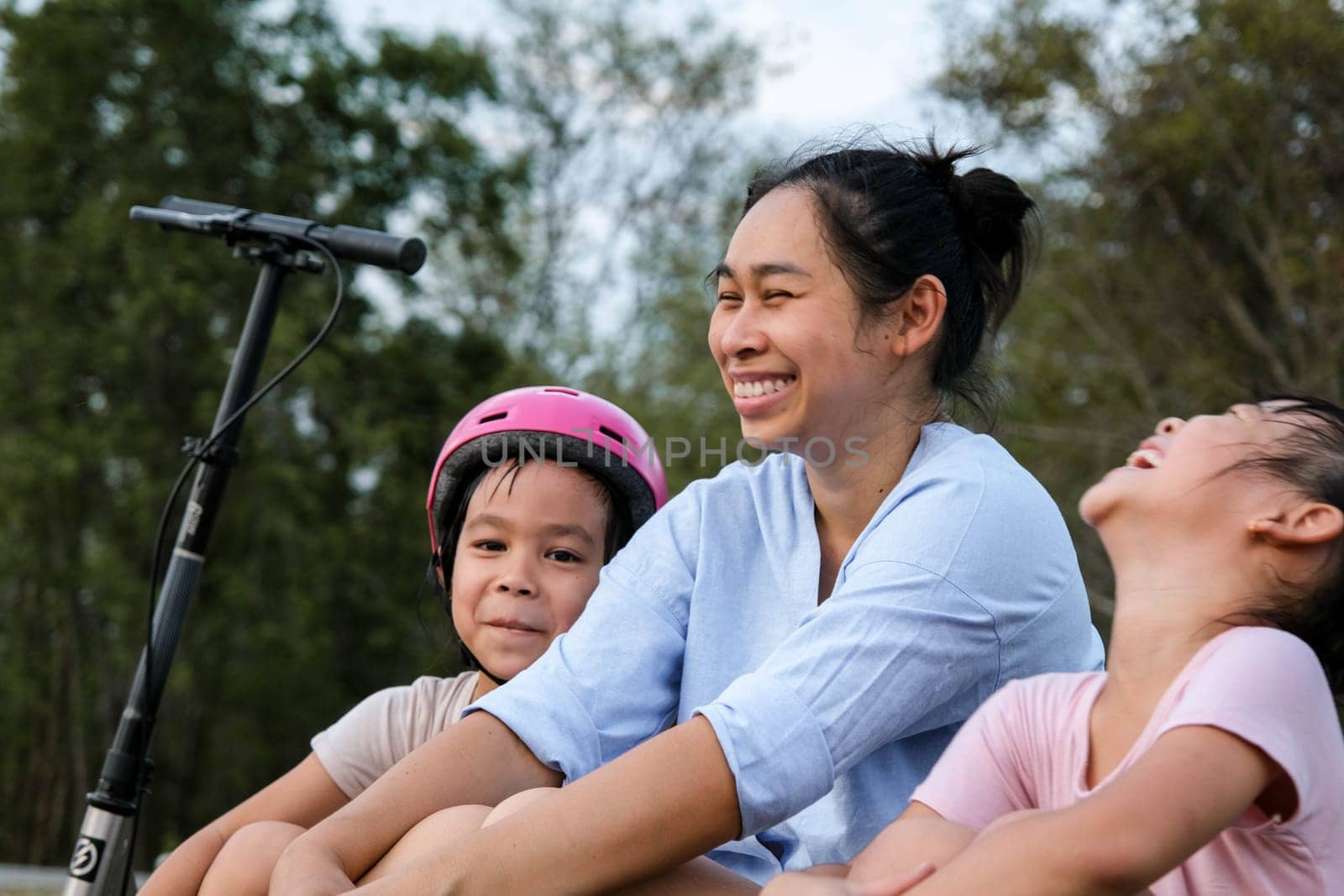 Mother and children sit and rest after riding scooters in the park. Mother and daughters spend their free time riding scooters outdoors together. Happy Loving Family. by TEERASAK