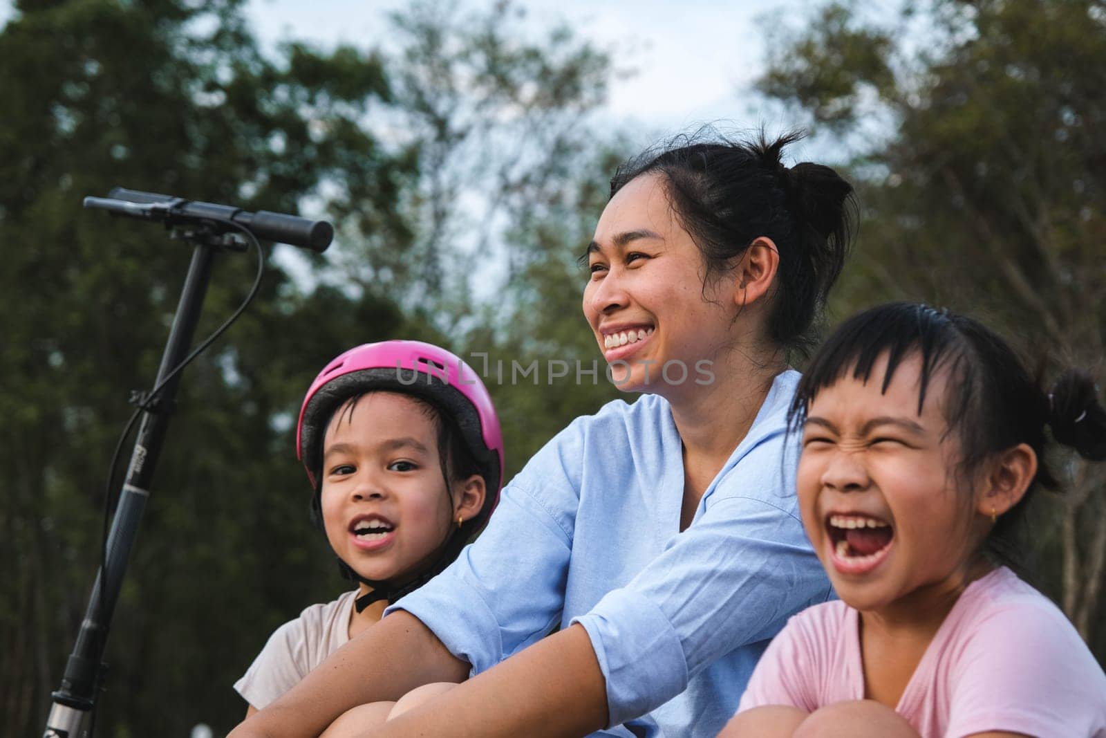 Mother and children sit and rest after riding scooters in the park. Mother and daughters spend their free time riding scooters outdoors together. Happy Loving Family.