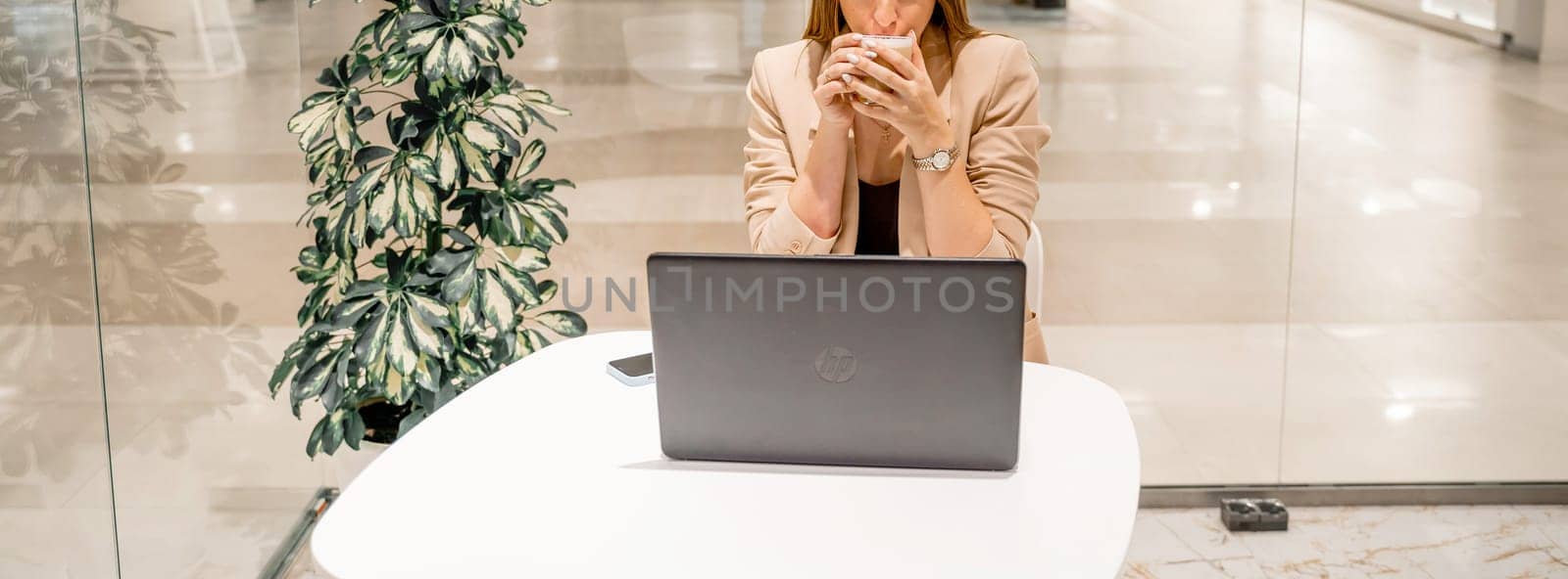 A business woman sits in a cafe, works at a computer, drinks coffee. She is wearing a beige jacket, brown top and black trousers. by Matiunina