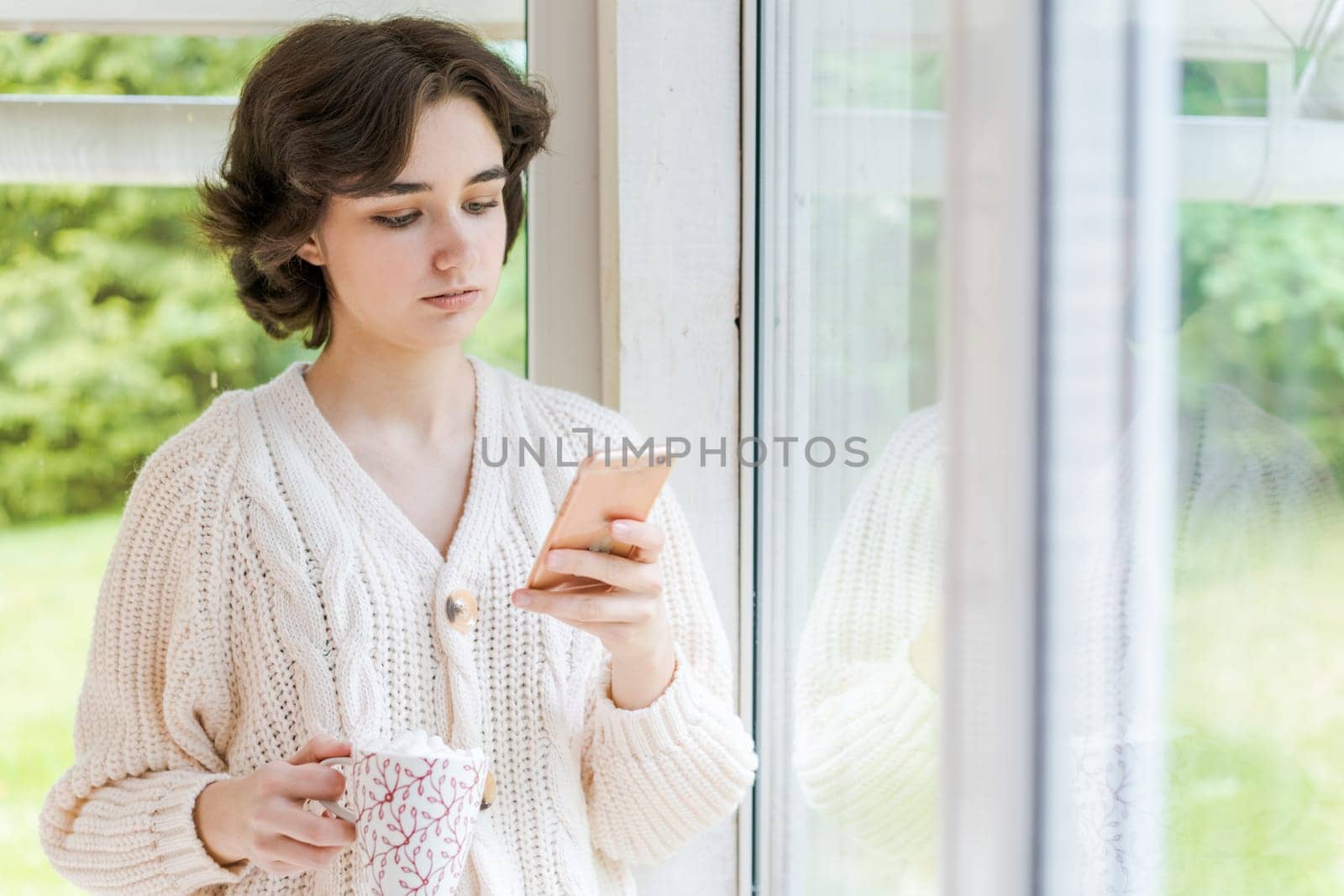 Portrait lonely caucasian young woman sitting near window with phone in her hand in a white knitted sweater, spending time in the morning on the veranda in a country house