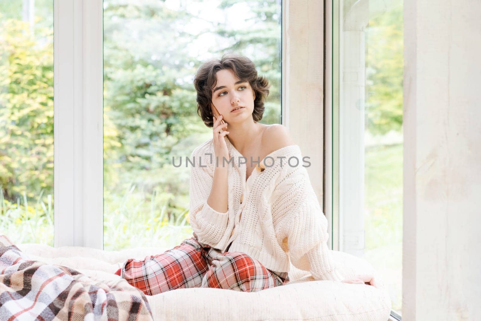 Portrait lonely caucasian young woman sitting near window with phone in her hand in a white knitted sweater, spending time in the morning on the veranda in a country house