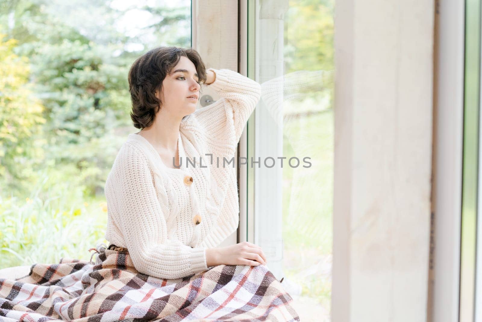 Portrait lonely caucasian young woman sitting near window apartments in a white sweater and a red plaid, spends time in the morning on the veranda in a country house