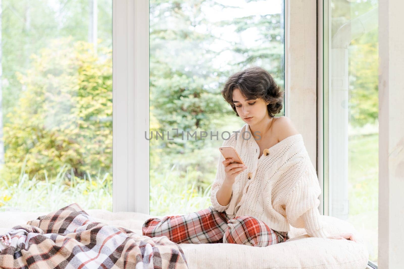 Portrait lonely caucasian young woman sitting near window with phone in her hand in a white knitted sweater, spending time in the morning on the veranda in a country house