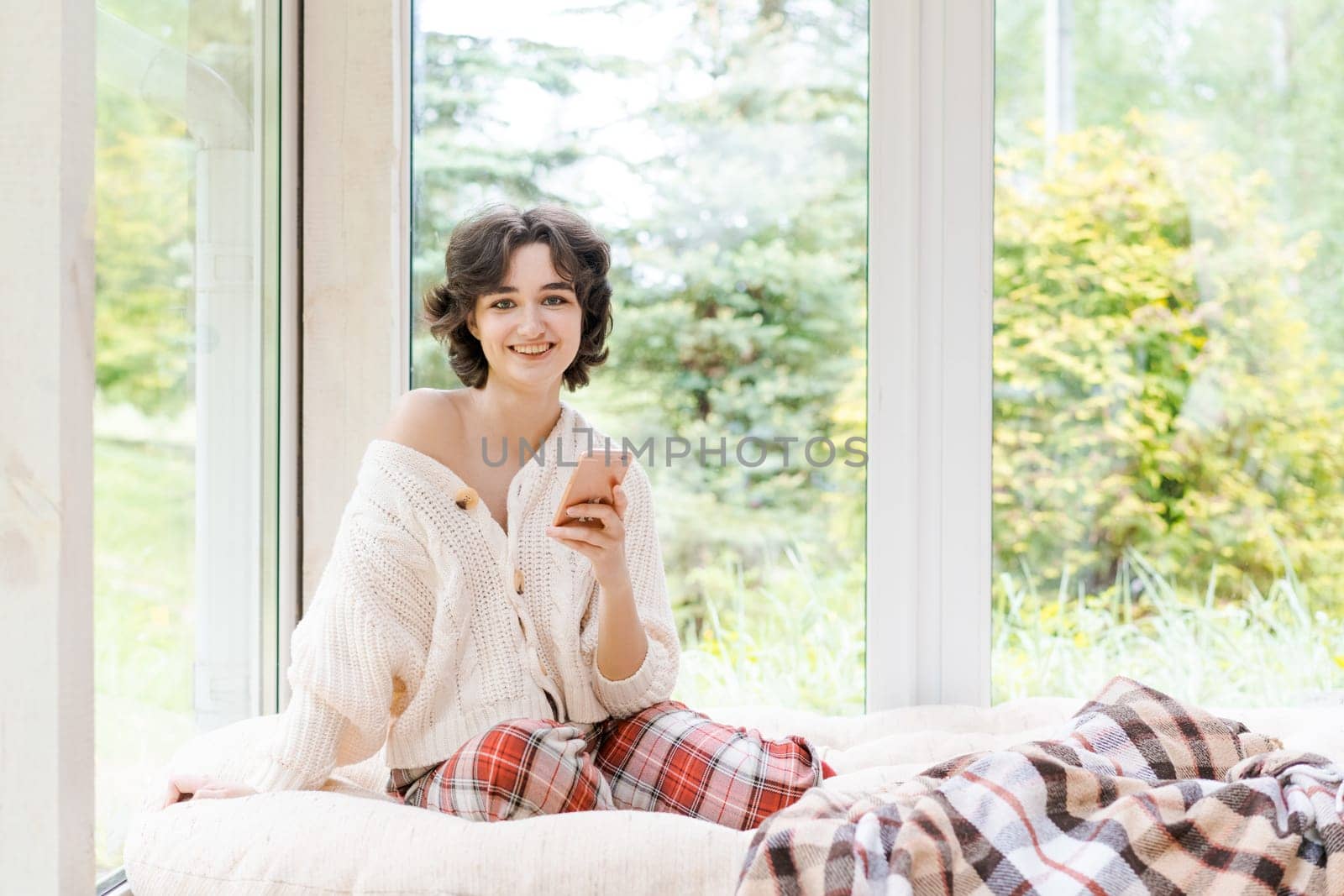 Portrait lonely caucasian young woman sitting near window with phone in her hand in a white knitted sweater, spending time in the morning on the veranda in a country house