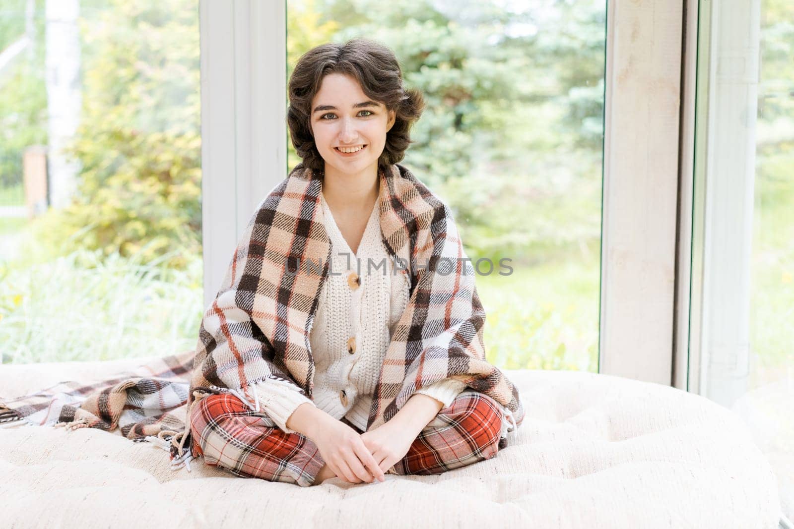 Portrait lonely caucasian young woman sitting near window apartments in a white sweater and a red plaid, spends time in the morning on the veranda in a country house