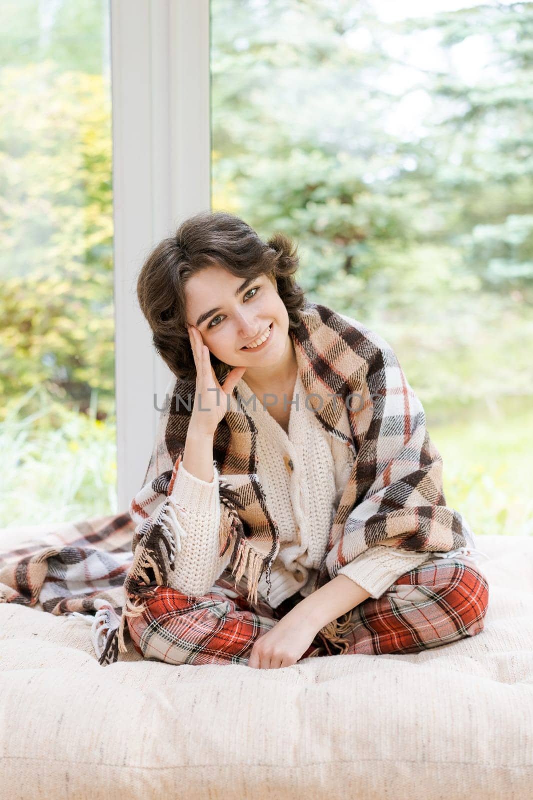 Portrait lonely caucasian young woman sitting near window apartments in a white sweater and a red plaid, spends time in the morning on the veranda in a country house