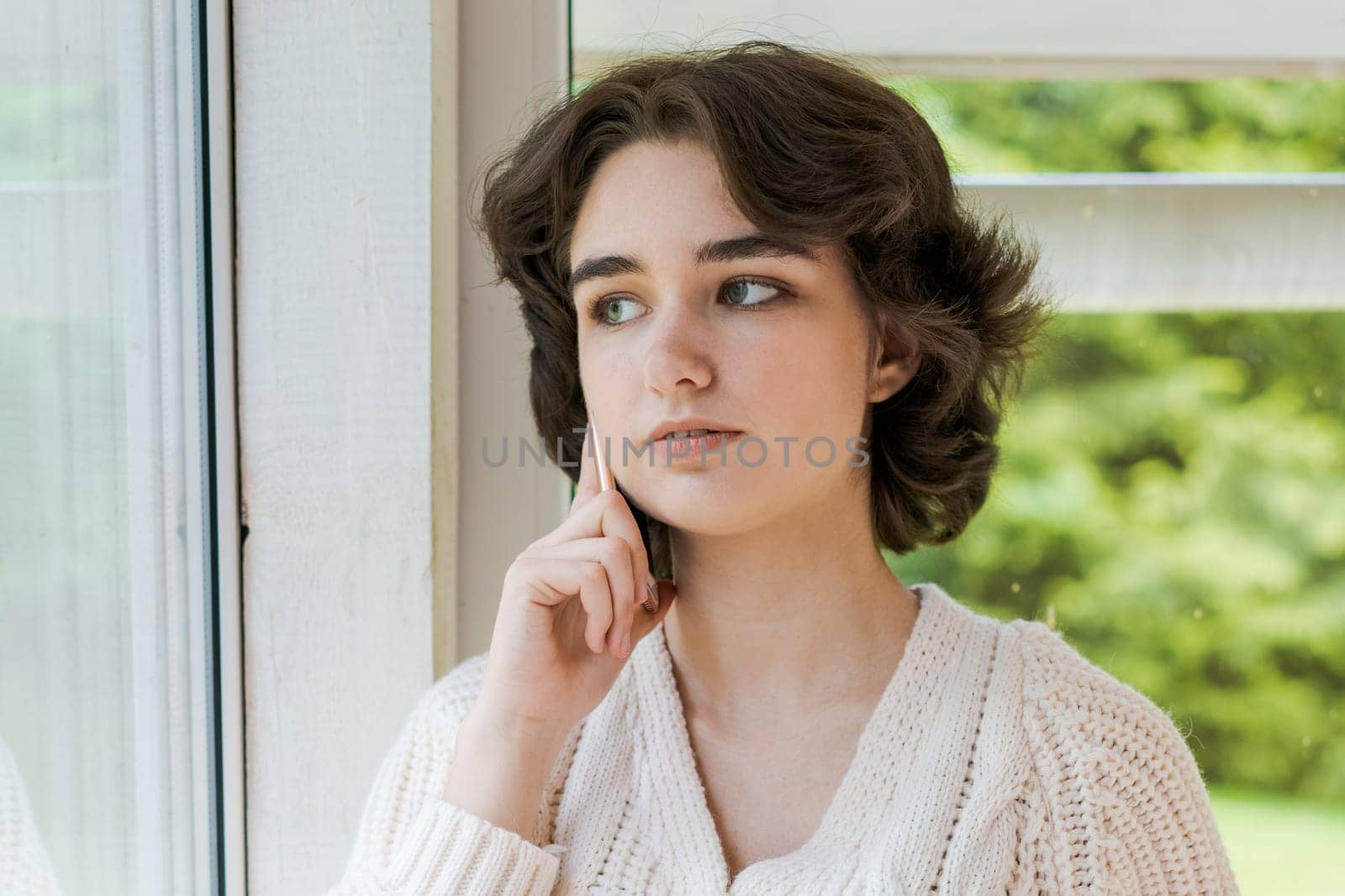 Portrait lonely caucasian young woman sitting near window with phone in her hand in a white knitted sweater, spending time in the morning on the veranda in a country house