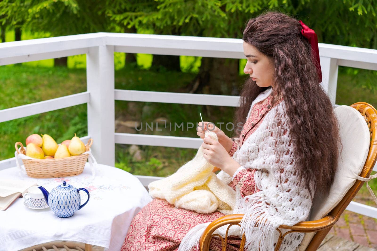 Young woman sitting on the terrace in backyard house sits in an armchair and knits, during the day against the background of trees in the garden