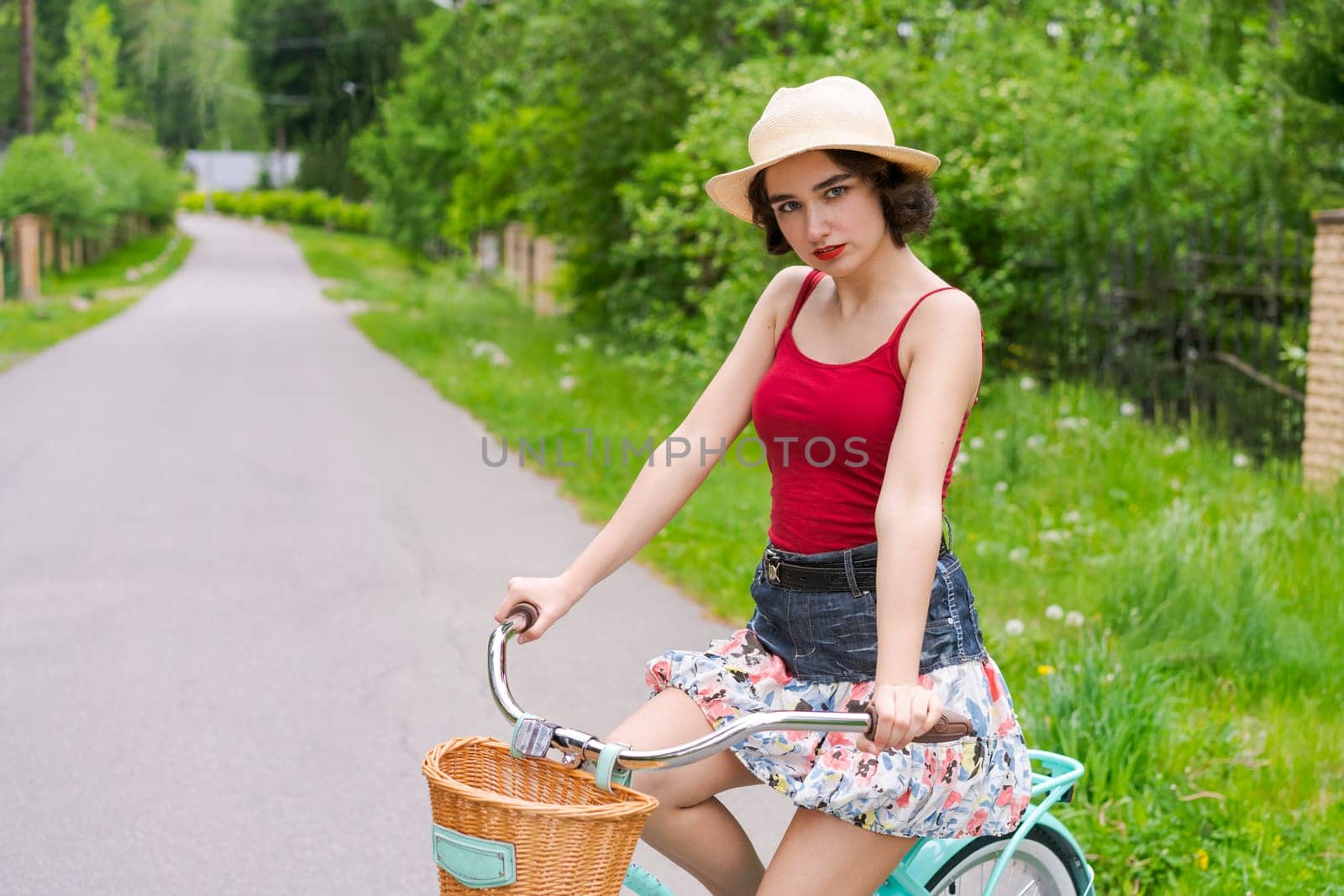 Portrait beautiful young girl in hat with bicycle on street in countryside in sunlight outdoor in a red tank top on a summer day rides on the weekend