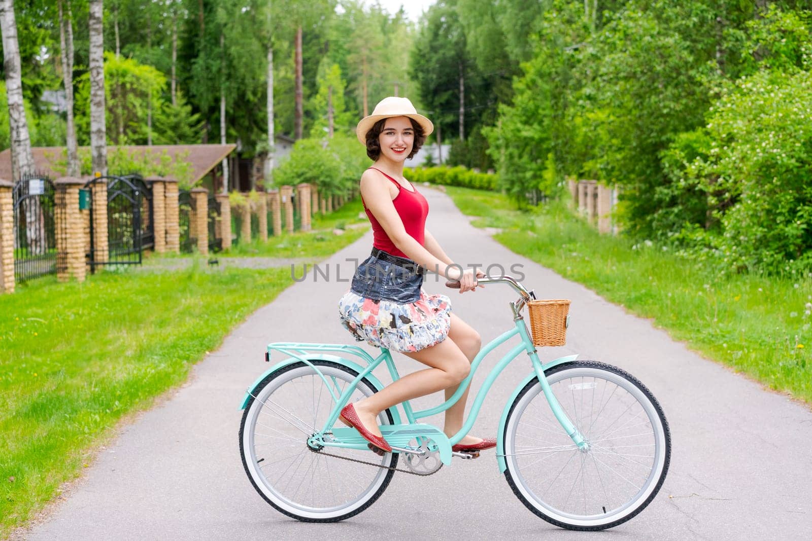 Portrait beautiful young girl in hat with bicycle on street in countryside in sunlight outdoor in a red tank top on a summer day rides on the weekend