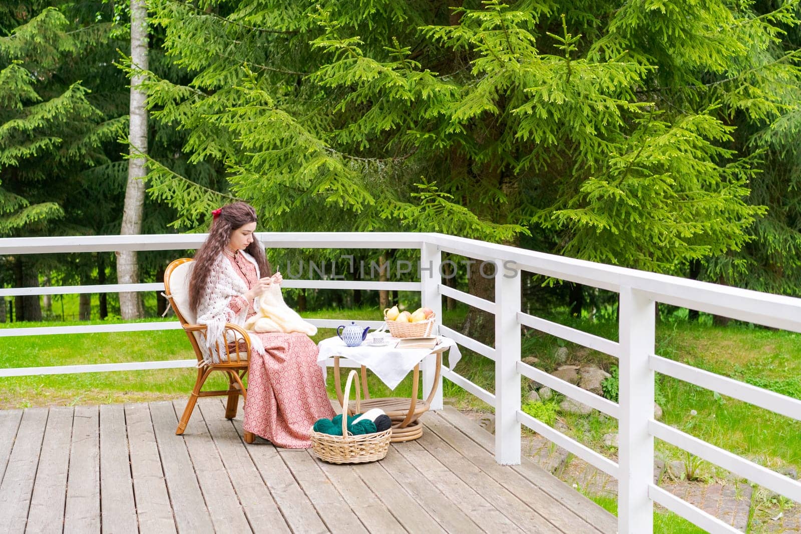 Young woman sitting on the terrace in backyard house sits in an armchair and knits, during the day against the background of trees in the garden