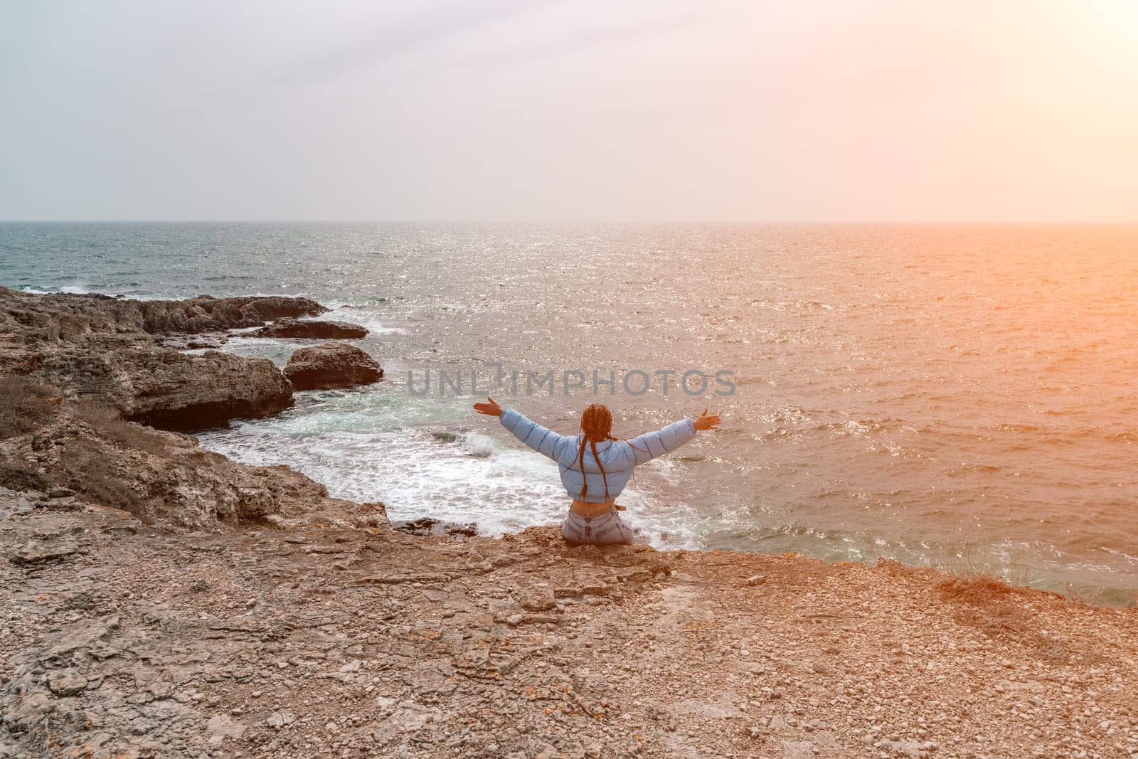 woman sea travel. A woman in a blue jacket sits on a rock above a cliff above the sea, looking at the stormy ocean. Girl traveler rests, thinks, dreams, enjoys nature.