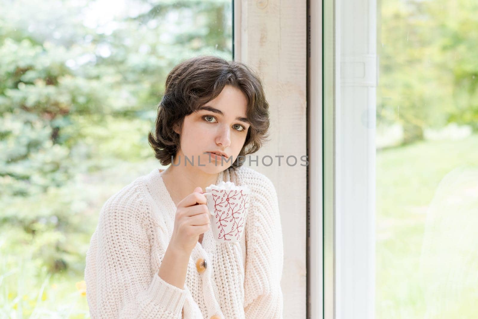 Female portrait on background window. Young woman sitting on veranda drinking her hot coffee. Cozy winter morning. Peace of mind and mental health. In a bright knitted sweater