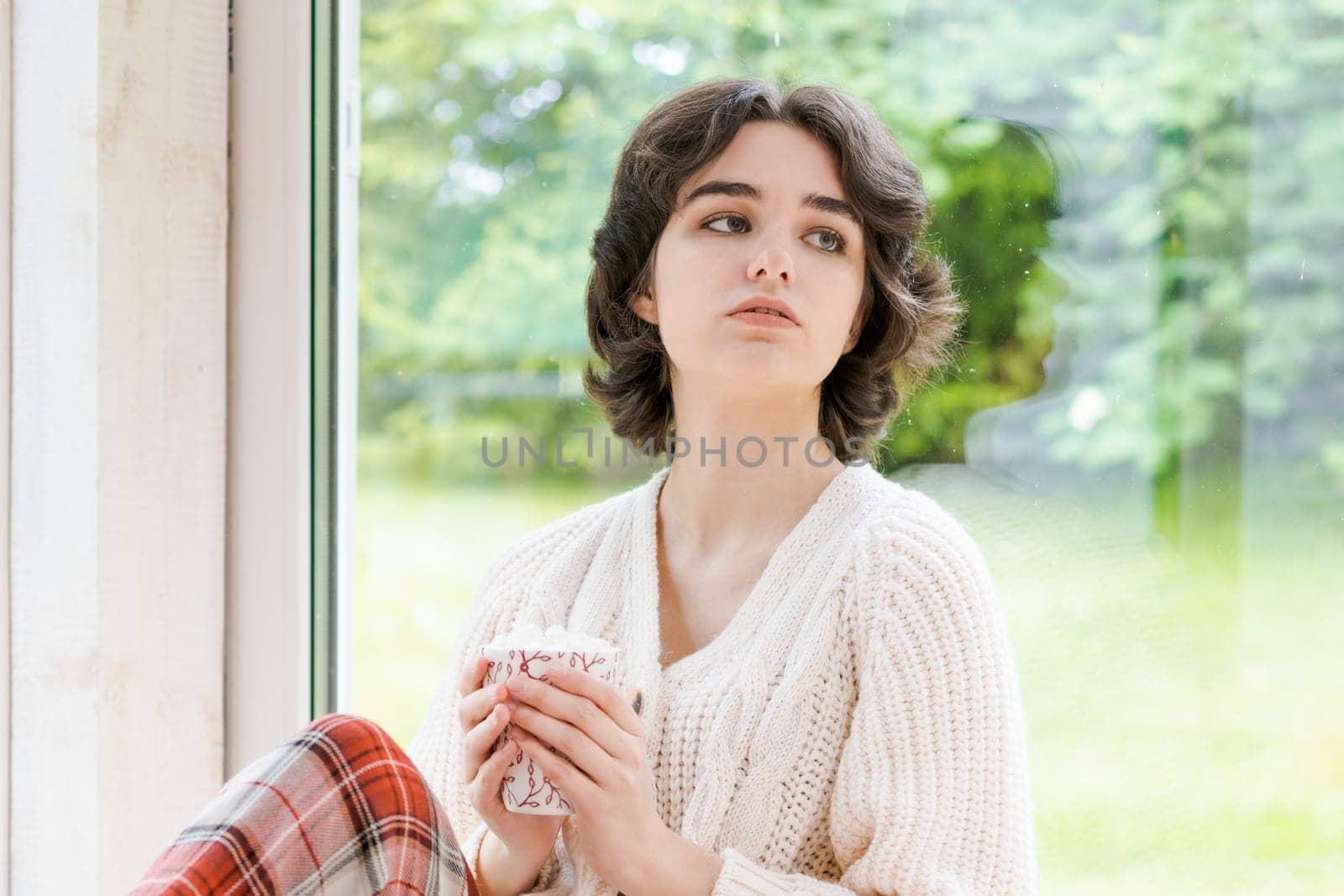 Female portrait on background window. Young woman sitting on veranda drinking her hot coffee. Cozy winter morning. Peace of mind and mental health. In a bright knitted sweater