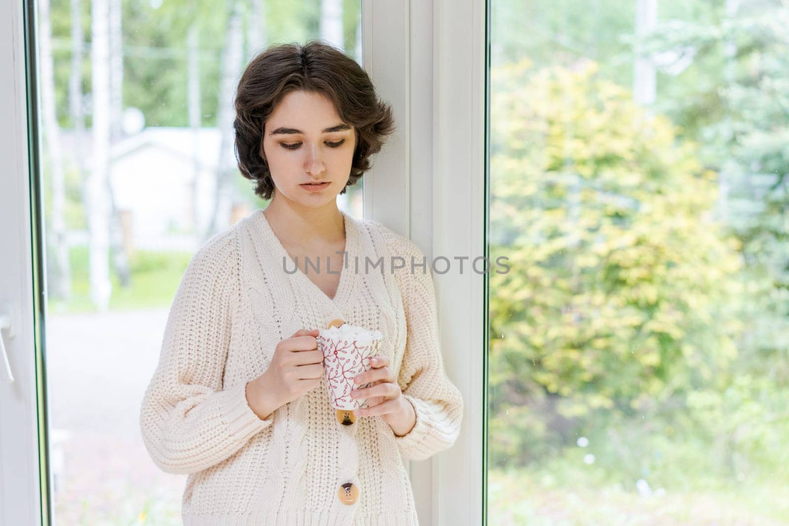 Female portrait on background window. Young woman sitting on the veranda drinking her hot coffee. Cozy winter morning. Peace of mind and mental health. In a bright knitted sweater