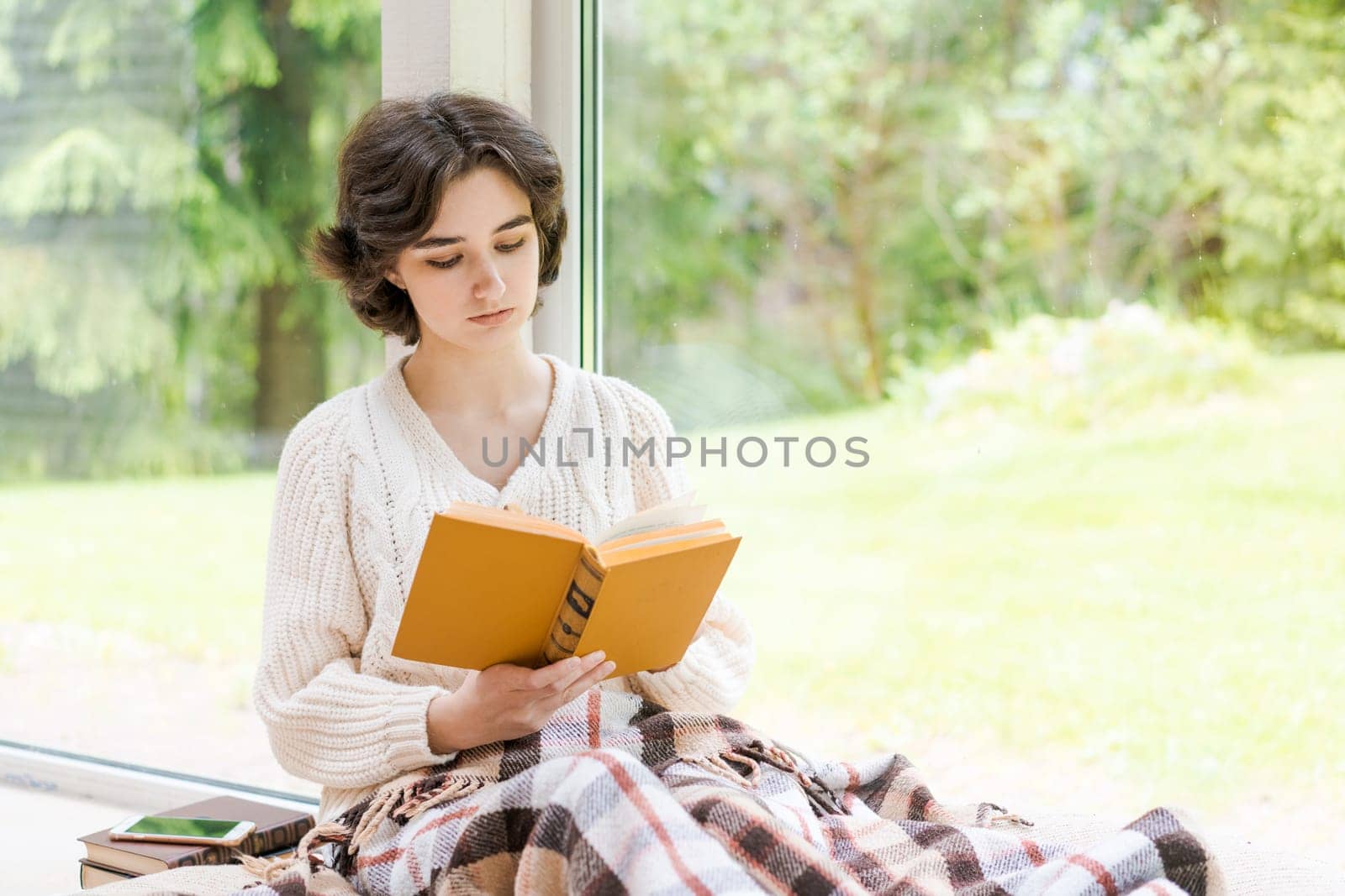 Brunette woman in warm sweater reading book sitting on room on the veranda by the window looking mood