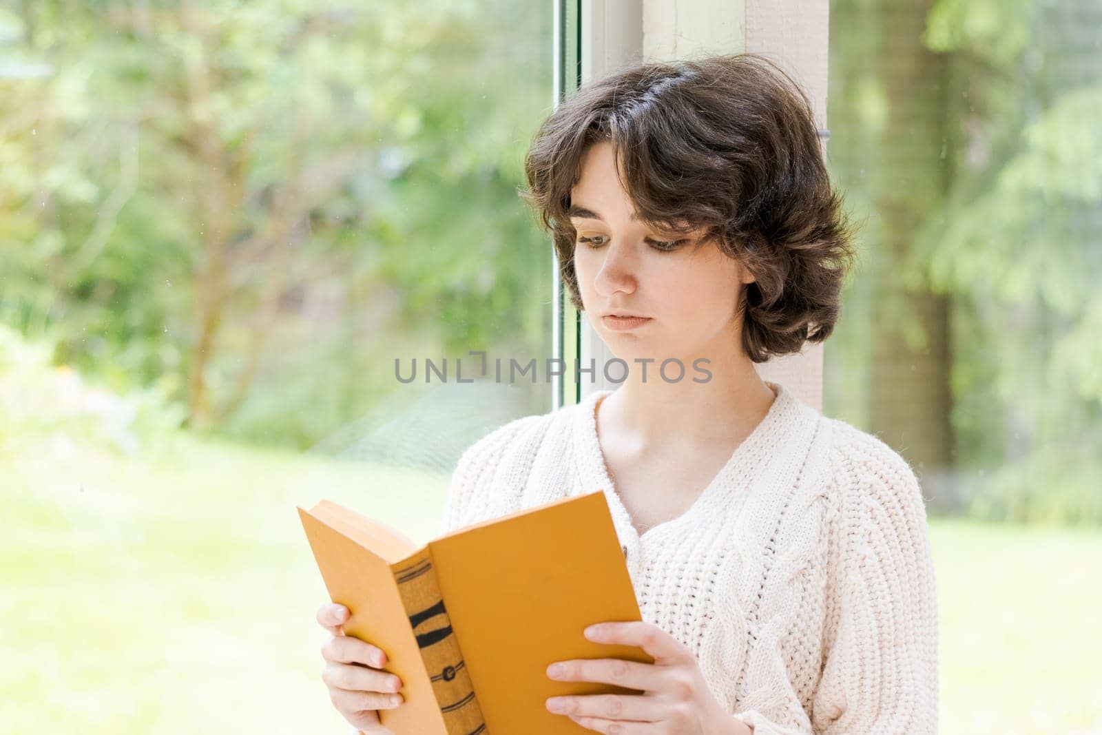 Brunette woman in warm sweater reading book sitting on room on the veranda by the window looking mood