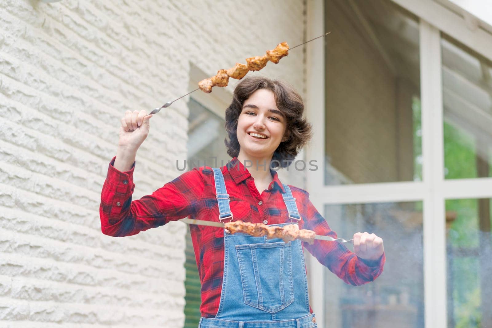 Barbeque party in garden with young woman in denim overalls and red plaid shirt in a country house on the terrace preparing meat skewers