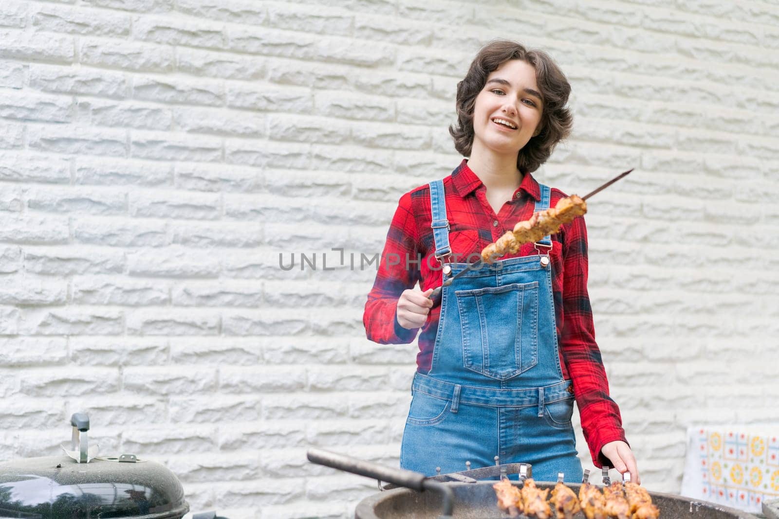 Barbeque party in garden with young woman in denim overalls and red plaid shirt by EkaterinaPereslavtseva