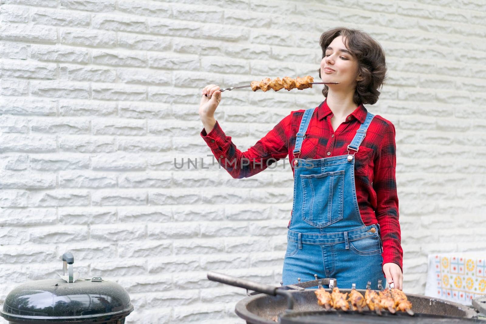 Barbeque party in garden with young woman in denim overalls and red plaid shirt in a country house on the terrace preparing meat skewers