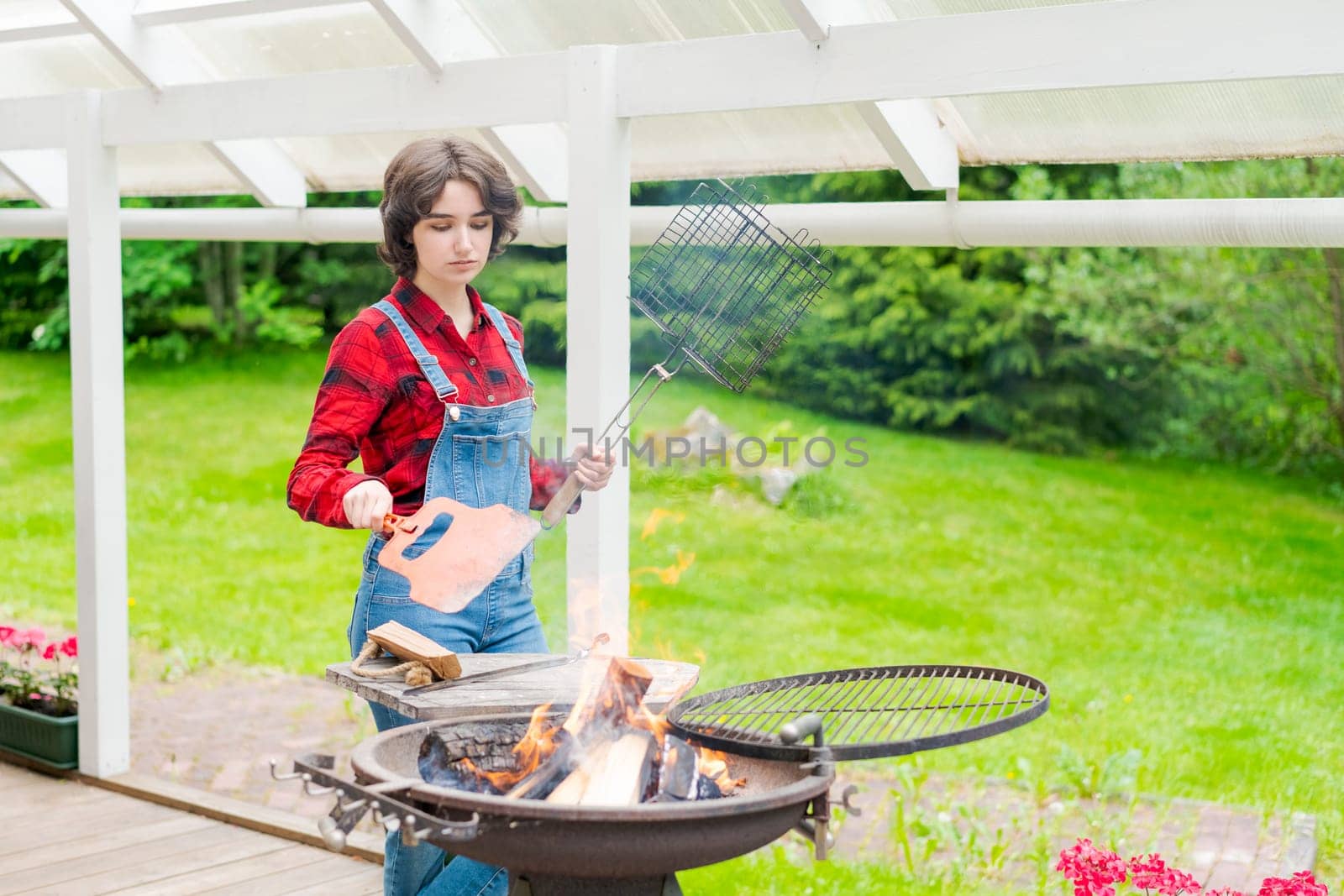 Barbeque party in garden with young woman in denim overalls and red plaid shirt in a country house on the terrace preparing meat skewers