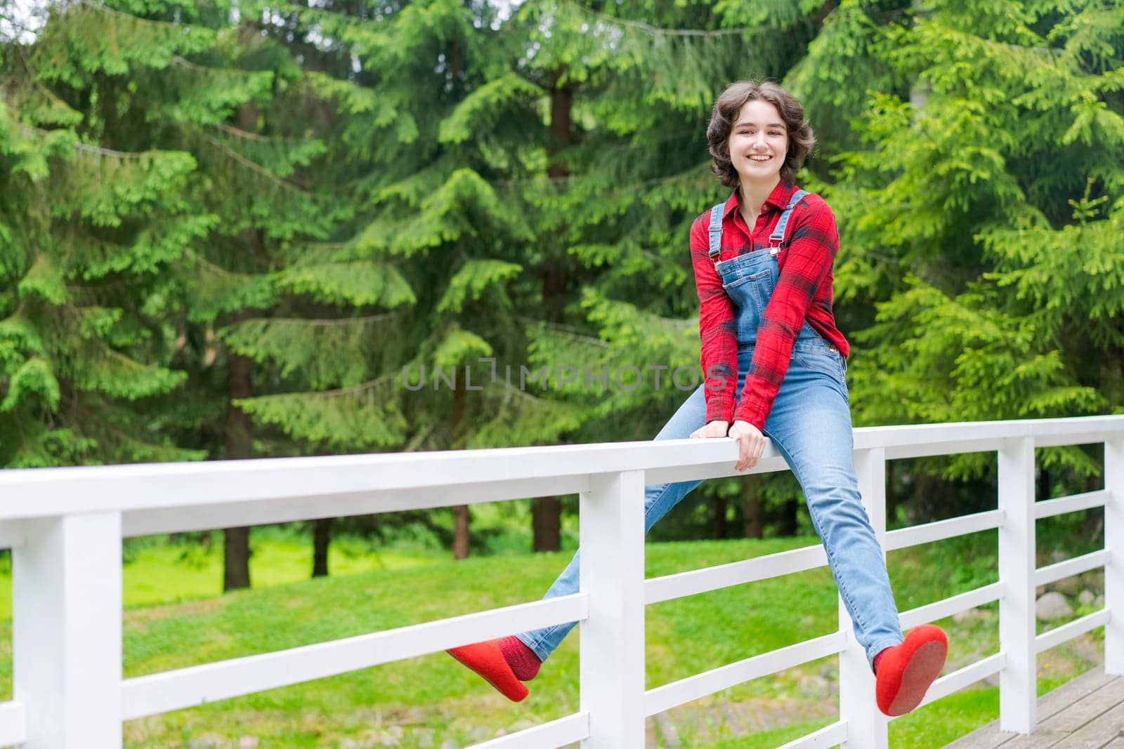 Happy young woman in blue denim overalls and red shirt sits on wooden fence on the terrace in the backyard, against the backdrop of a green lawn and forest on a sunny day