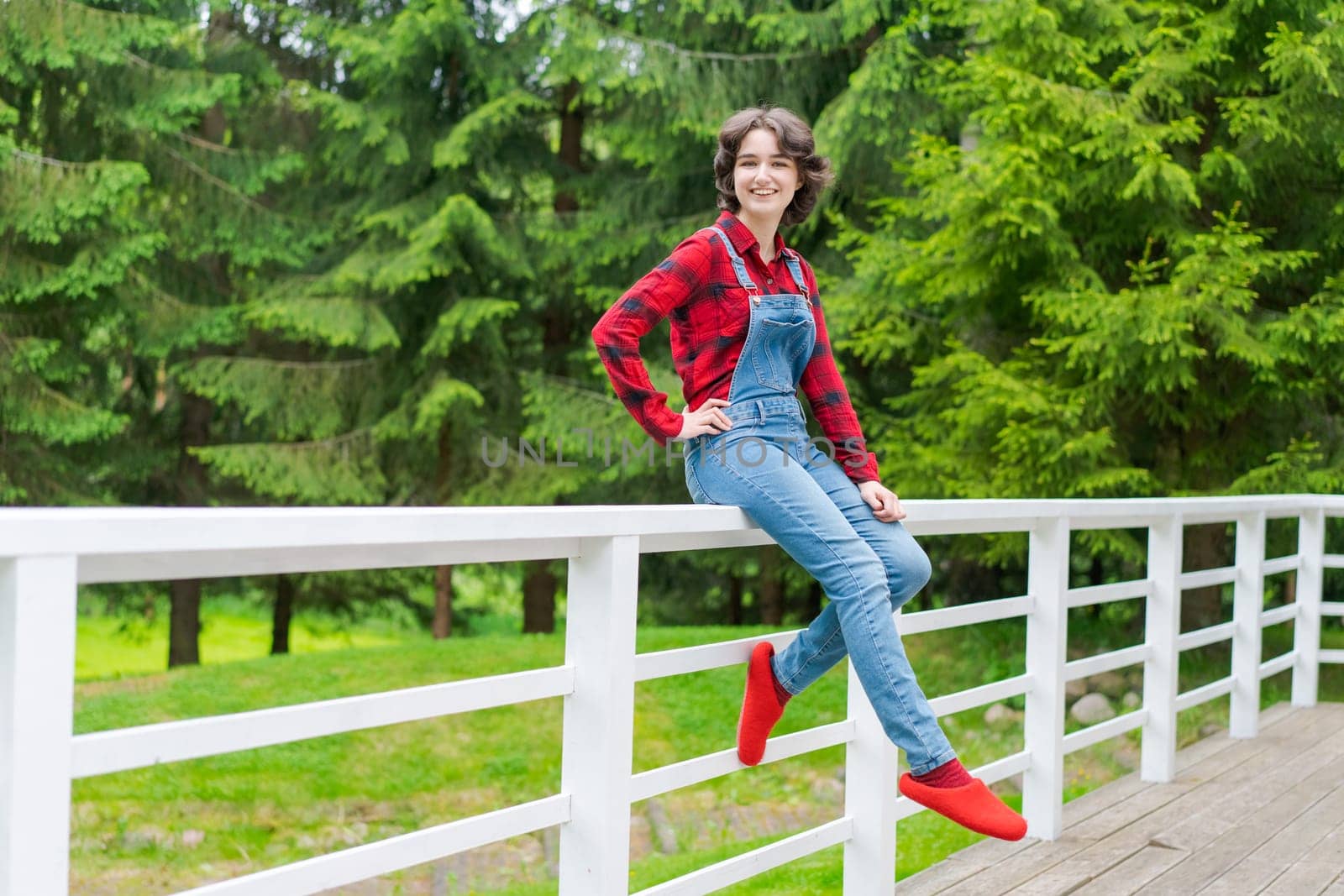 Happy young woman in blue denim overalls and red shirt sits on wooden fence by EkaterinaPereslavtseva