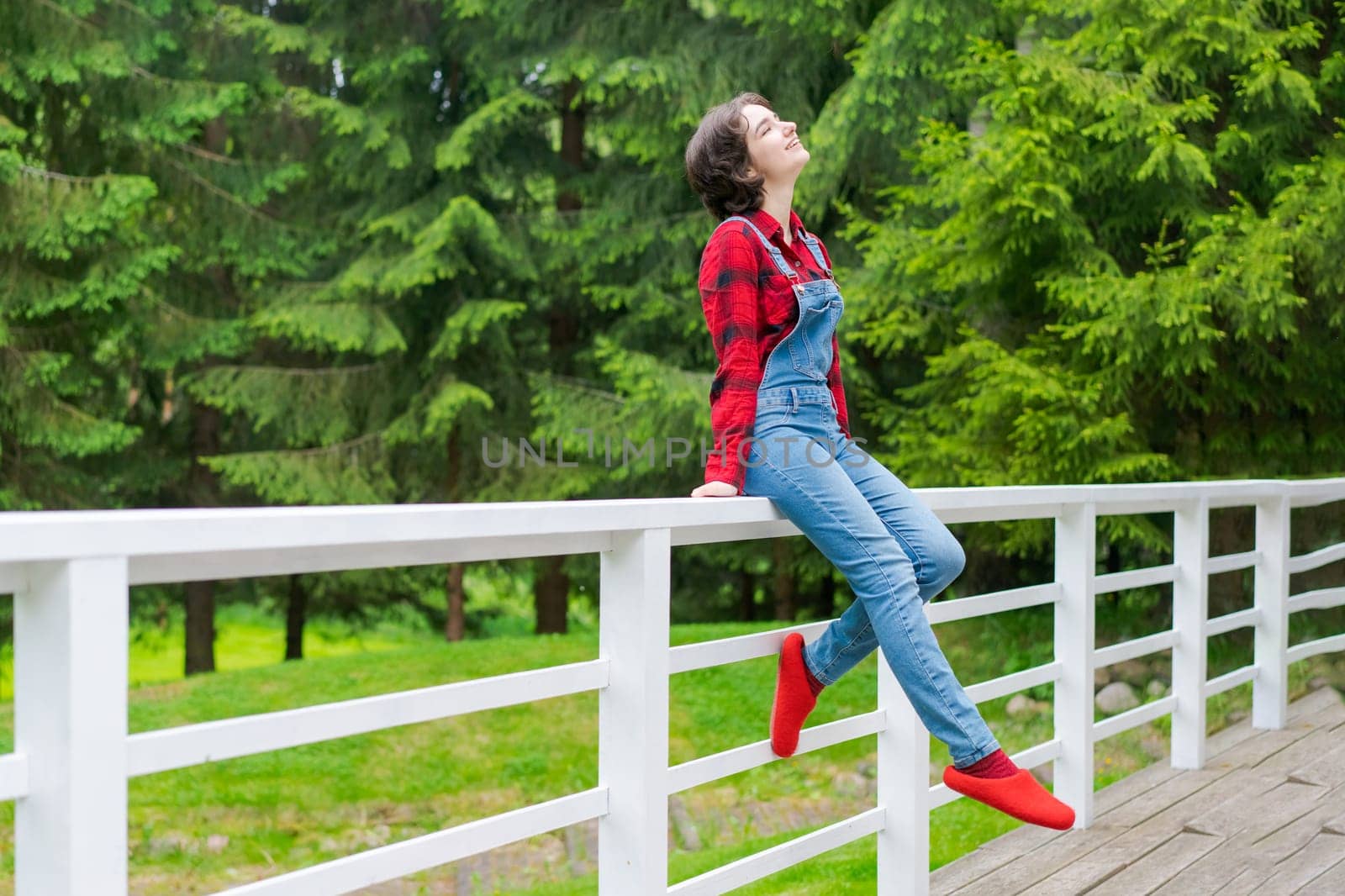 Happy young woman in blue denim overalls and red shirt sits on wooden fence on the terrace in the backyard, against the backdrop of a green lawn and forest on a sunny day