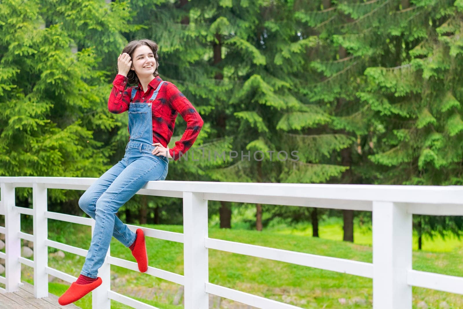 Happy young woman in blue denim overalls and red shirt sits on wooden fence on the terrace in the backyard, against the backdrop of a green lawn and forest on a sunny day