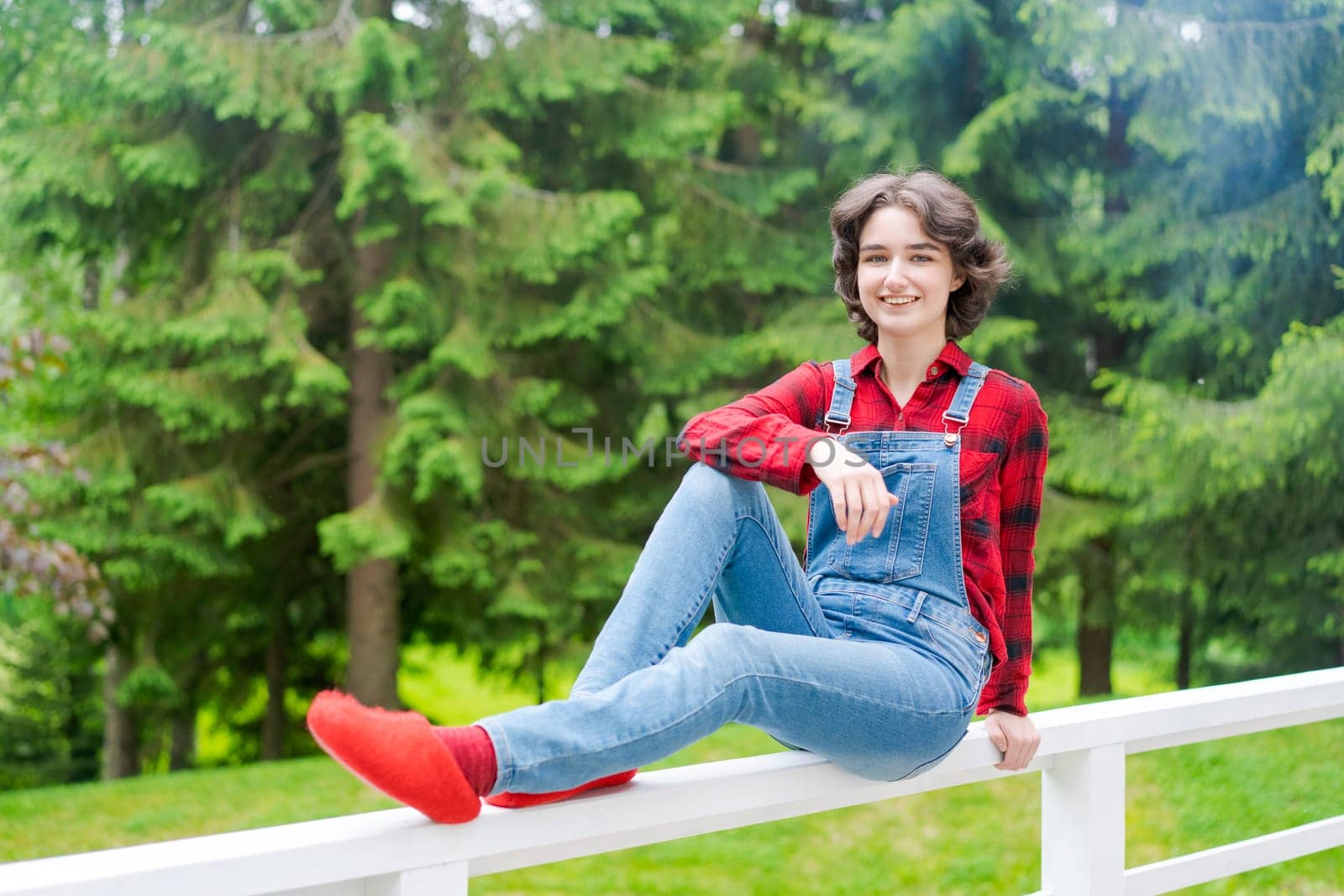 Happy young woman in blue denim overalls and red shirt sits on wooden fence on the terrace in the backyard, against the backdrop of a green lawn and forest on a sunny day